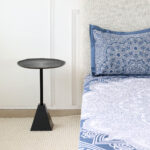 Modern black metal side table with textured round top and pyramid base placed beside a bed
