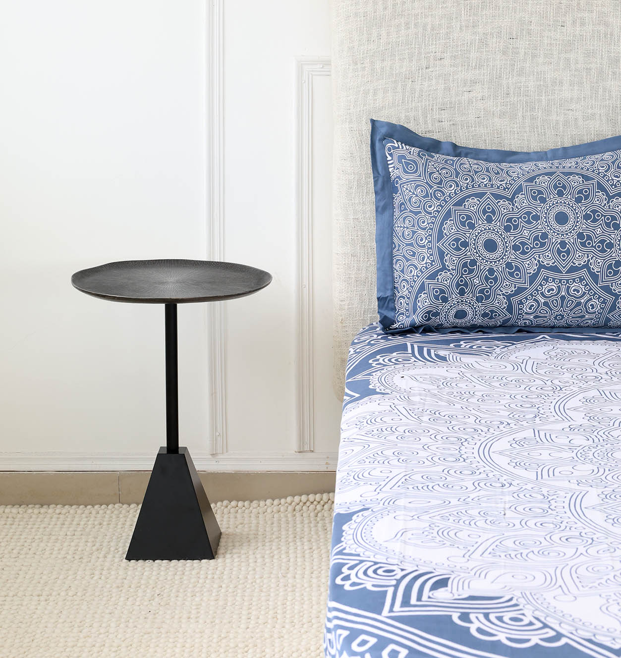Black Metal Side Table – Round Textured Top with Pyramid Base Beside Bed | Modern Industrial Design Modern black metal side table with textured round top and pyramid base placed beside a bed