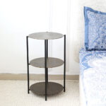Round wooden side table with black metal base placed beside the bed – modern bedside furniture