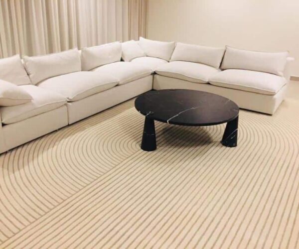 Rectangular radial pattern rug in light beige with fine repeating concentric stripe lines, grounding a white sectional sofa and black marble coffee table.