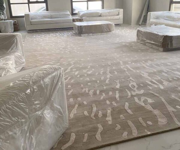 Large beige minimalist textured area rug with a low-relief, sand ripple pattern, providing seamless floor coverage in a bright, modern room with neutral curtains.