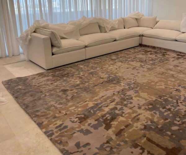 Large beige minimalist textured area rug with a low-relief, sand ripple pattern, providing expansive coverage in a modern room with sheer curtains.