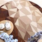 An Earth Tone Geometric Pattern Round Area Rug with a faceted triangular pattern in brown, taupe, and beige tones styled with a wooden side table.