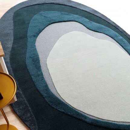 A Layered Abstract Shaped Rug with concentric organic shapes in teal, charcoal, and light grey placed on a wooden floor.