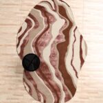 A Wool Shaped Rug with an irregular organic silhouette featuring rhythmic wavy patterns in brown, beige, and rose.